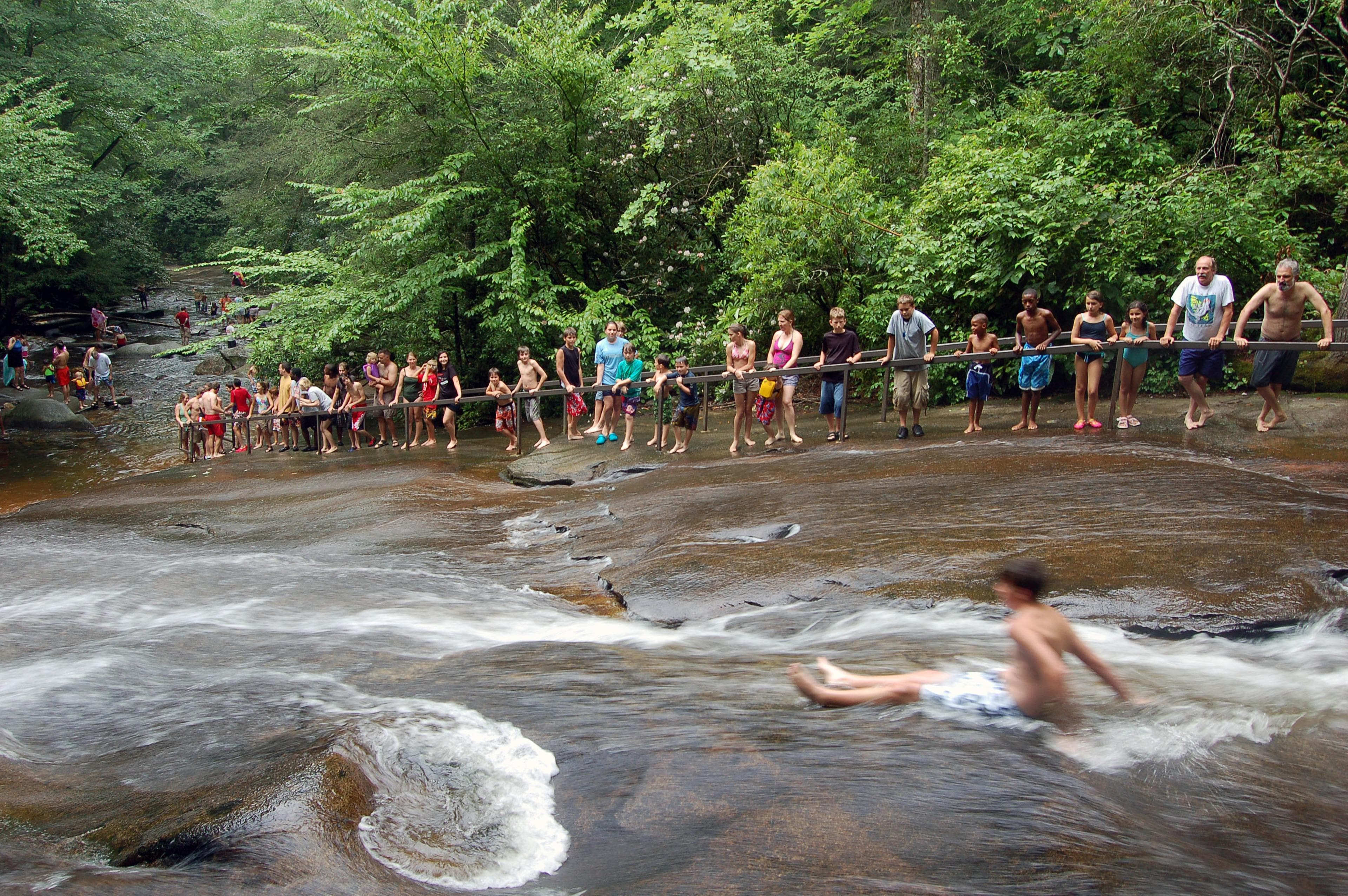A person slides down the natural waterfall in front of a line of people with green trees in the background A person slides down the natural waterfall in front of a line of people with green trees in the background