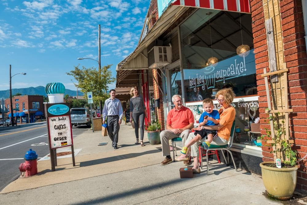 Downtown exterior shot of Black Mountain's hey hey cupcake! local bakery, with a young couple walking in the background and an elderly couple with their grandchild sitting in chairs in the foreground. Downtown exterior shot of Black Mountain's hey hey cupcake! local bakery, with a young couple walking in the background and an elderly couple with their grandchild sitting in chairs in the foreground.