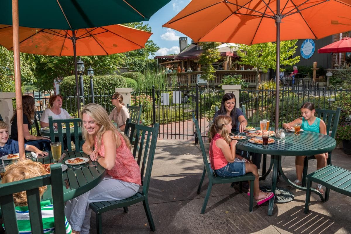 Three patio tables with colorful umbrellas each have people seated and enjoying food in the foreground, with lush landscaping and distant businesses in the background. Three patio tables with colorful umbrellas each have people seated and enjoying food in the foreground, with lush landscaping and distant businesses in the background.