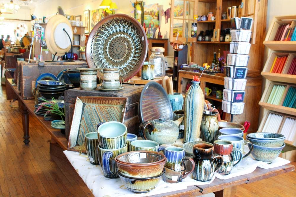 Interior image of a gift shop with warm color theme - a table with locally crafted pottery is in the foreground, with cards, books, and other gifts in the background. Interior image of a gift shop with warm color theme - a table with locally crafted pottery is in the foreground, with cards, books, and other gifts in the background.