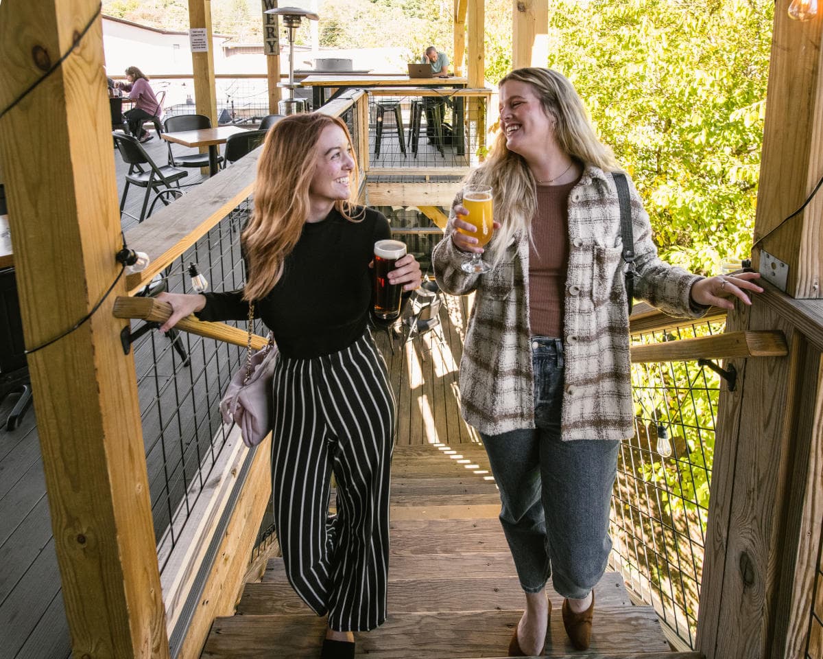 Two people with long hair climb a flight of stairs that leads to an outdoor patio, each holding a beer. Two people with long hair climb a flight of stairs that leads to an outdoor patio, each holding a beer.